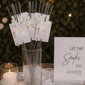 Op de afbeelding: Een heldere glazen vaas gevuld met grijze sterretjes, elk bevestigd aan een witte kaart met gouden tekst Let Sparks Fly, Emma & James, en 05.01.2027. Een wit bordje in de buurt toont de tekst Let The Sparks Fly! Let's light the way.