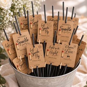 May include: A collection of sparklers in a metal bucket, each attached to a rectangular kraft paper tag. The tags feature black text including phrases like Let Love Sparkle, Let Sparks Fly, and Cheers to Love, along with the names Emma & James and the date 05.01.2027. Each tag holds a single matchstick.