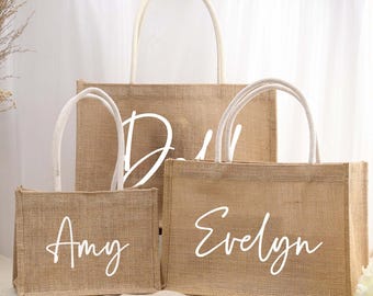 May include: Three jute tote bags with white handles and personalized names in white script: "Amy", "Evelyn", and "D". The bags are in varying sizes, with a natural tan color. The bags are likely for a wedding or event.