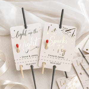 Op de afbeelding: Twee rechthoekige witte papieren labels met afgeronde hoeken, elk met een zwarte sterretje en een lucifer. Het linker label heeft zilveren tekst met light up the Night, Emma & James, 05.01.2027. Het rechter label heeft gouden tekst met Let Sparks Fly, Emma & James, 05.01.2027.