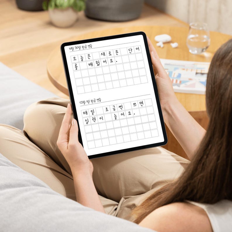 May include: A black tablet displaying a Korean language practice sheet with grid boxes for writing. The tablet is held by a person, with text visible in Korean. The tablet is on a person's lap.