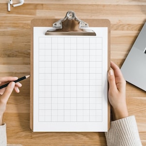 May include: A wooden clipboard with a silver clip holds a grid-lined white paper. A hand holds a black pencil, poised to write. A laptop and a pair of white earbuds are visible on the wooden surface.