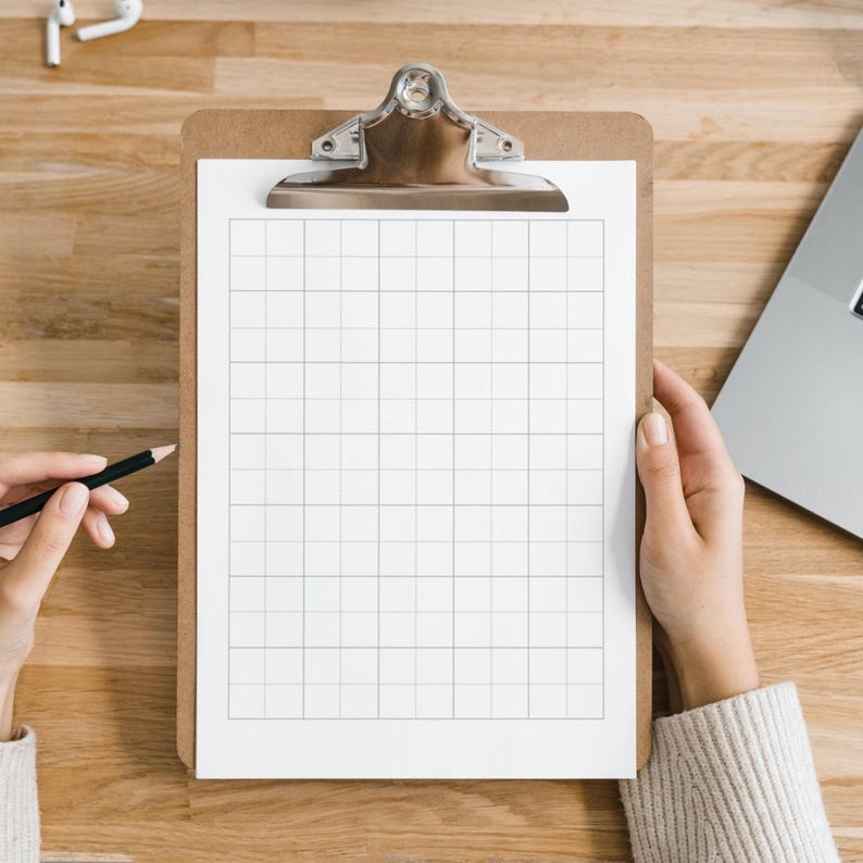 May include: A wooden clipboard with a silver clip holds a grid-lined white paper. A hand holds a black pencil, poised to write. A laptop and a pair of white earbuds are visible on the wooden surface.