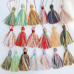 May include: A collection of colorful handmade tassels with various color combinations. Each tassel features a looped top and a long, fringed bottom made of multiple strands of thread. The tassels are in shades of green, red, yellow, purple, and pink.