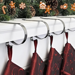 Può includere: Una fila di porta calze in metallo argentato attaccati a una mensola bianca, con calze di Natale rosse appese sotto. La mensola è decorata con una ghirlanda verde e biscotti di pan di zenzero.