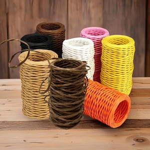 Pode incluir: Uma coleção de rolos de barbante coloridos em várias tonalidades, incluindo marrom, preto, bege, branco, rosa, amarelo e laranja. O barbante é bem enrolado, e alguns rolos têm fios soltos. Os rolos estão dispostos sobre uma superfície de madeira.