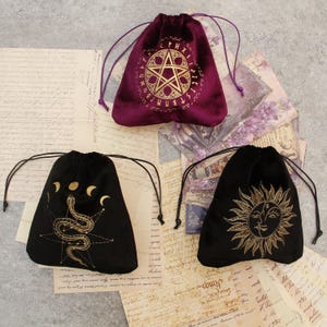 Può includere: Tre sacchetti in velluto con coulisse in bordeaux e nero. La borsa bordeaux presenta un design a pentagramma dorato. Le borse nere hanno disegni dorati di un serpente, mezzelune e un sole con un volto. Ogni borsa ha una chiusura con coulisse.