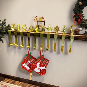 May include: A Christmas stocking holder with gold-colored hooks and decorative ornaments. Two red Christmas stockings with snowman and reindeer designs hang from the hooks. The ornaments include a bow, wreath, boot, gift, tree, and snowflake.