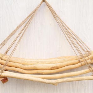 Puede incluir: Estante colgante de madera natural clara y cuerda. El estante se suspende de un solo punto, con múltiples hebras de cuerda que sostienen la base de madera. Las ramas están dispuestas horizontalmente, creando un diseño rústico y minimalista.