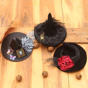 May include: Three miniature black witch hats decorated with distinct embellishments. One features a silver cameo and butterflies, another has a gold spider charm and black lace, and the third is adorned with a large red fabric rose and black netting.