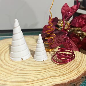 May include: Two white, cone-shaped ornaments with spiral grooves sit on a wooden slice. Beside them are two decorative wire spirals, one gold and one red, and a red flower. The ornaments are likely for holiday decor.