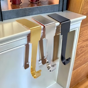 May include: Three metal stocking holders in gold, silver, and black hang from a white mantelpiece. The holders have a flat top that rests on the mantel and a curved hook to hold stockings or other decorations. The mantelpiece is white with decorative moulding.