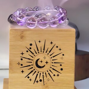 Op de afbeelding: Een armband van doorschijnende paarse bolvormige kralen rustend op een lichtgekleurde houten kubus. De kubus is voorzien van een lasergegraveerd ontwerp van een maansikkel omringd door stralende zonnestralen, stippen en kleine vierpuntige sterren.