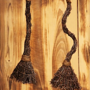 May include: Two rustic, handmade miniature brooms hang against a wooden background. The brooms are crafted from dark brown twigs and bound with twine. They are suspended by thin, natural-colored cords, perfect for Halloween or rustic decor.