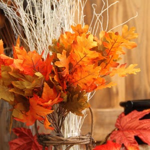 May include: A fall-themed arrangement featuring vibrant orange and yellow artificial oak leaves. The leaves are arranged in a clear glass jar with a woven basket base, complemented by white twigs. A decorative autumn display.