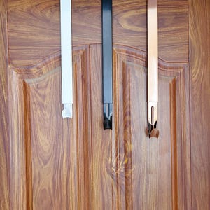 May include: Three over-the-door hooks in white, black, and rose gold hang from the top of a wooden door. Each hook has a rectangular, flat design with a curved hook at the bottom. The door has a wood grain pattern.