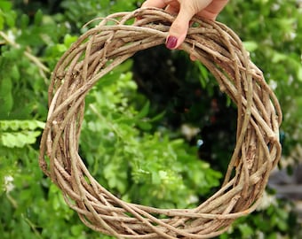 May include: A natural grapevine wreath, held by a hand with painted nails, against a backdrop of green foliage. The wreath is made of intertwined brown branches, creating a circular shape. Perfect for seasonal decor or crafting.