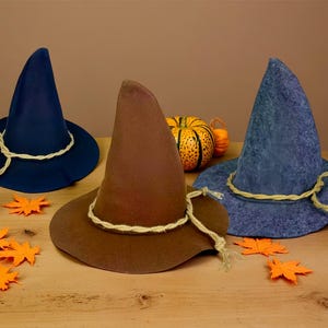 May include: Three whimsical witch hats in navy blue, brown, and blue-grey, adorned with rope trim. Miniature pumpkins and orange leaf cutouts decorate the wooden surface, creating a festive autumn scene.