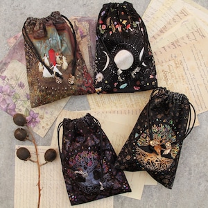 Puede incluir: Cuatro bolsas con cordón con diseños únicos. Una presenta una escena de bosque con figuras, otra tiene un diseño celestial con lunas y estrellas, y otras dos muestran motivos de árbol de la vida. Cada bolsa tiene un cierre de cordón negro y dijes decorativos.