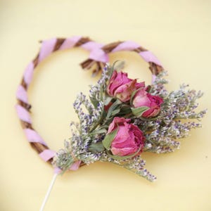 Könnte beinhalten: Ein Strauß getrockneter rosa Rosen und lila Blumen, die mit Schnur zusammengebunden und an einem Zweig-Herzenkranz befestigt sind.