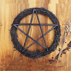 May include: A black pentagram wreath made of twigs and branches, with a five-pointed star in the centre. The wreath is set against a wooden background, with dried flowers to the right.