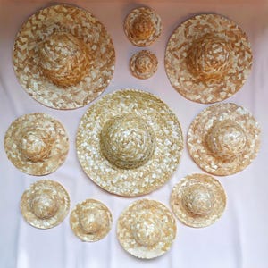 Puede incluir: Una colección de sombreros de paja tejidos de varios tamaños. Los sombreros son de color marrón claro natural y tienen un ala ancha. Los sombreros están dispuestos sobre un fondo rosa claro. Los sombreros son probablemente para protección solar o fines decorativos.