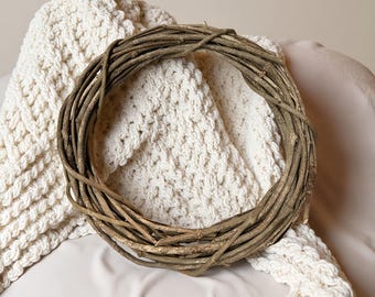 May include: A circular wreath made of intertwined brown twigs. The wreath is resting on a cream-colored, textured blanket. The blanket is draped over a light beige surface, creating a neutral backdrop for the wreath.