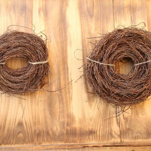 May include: Two brown twig wreaths, tied with twine, are displayed against a wooden background. The wreaths are circular, with a hole in the centre, and appear to be handmade. The wood has a warm, natural tone.