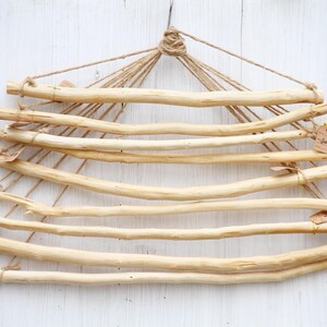 Op de afbeelding: Een natuurlijke houten wandhanger met meerdere horizontale deuvels die aan touw zijn opgehangen. De deuvels zijn lichtbruin en gerangschikt in een driehoekige vorm. Kleine labels zijn aan sommige deuvels bevestigd. Voor het weergeven van items.