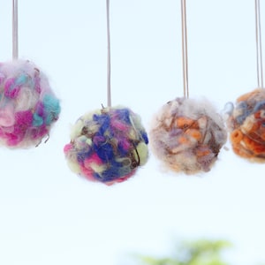 Pode incluir: Quatro bolas de fio coloridas penduradas. Cada bola apresenta uma mistura diferente de cores, incluindo rosa, azul, laranja e branco. As bolas de fio são suspensas por fios finos prateados contra um fundo azul claro.