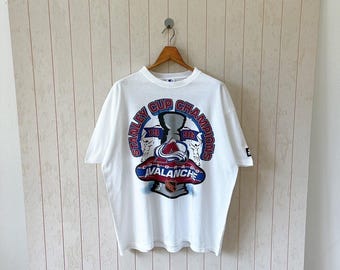 May include: White t-shirt featuring a graphic of the Colorado Avalanche hockey team. The design includes the words "Stanley Cup Champions" and the team's logo in blue, red, and silver. The shirt is hanging on a wooden hanger.