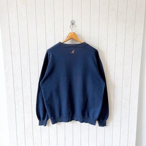 May include: Navy blue sweatshirt with a crew neck and long sleeves. A small, embroidered kangaroo logo is centered on the upper back. The sweatshirt is hanging on a wooden hanger against a white background.