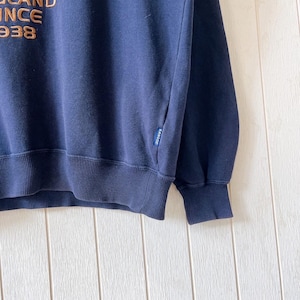 May include: Navy blue sweatshirt with ribbed cuffs and hem. The upper left features the text "BRAND SINCE 1938" in a gold colour. A small tag with the brand name is visible on the sleeve.