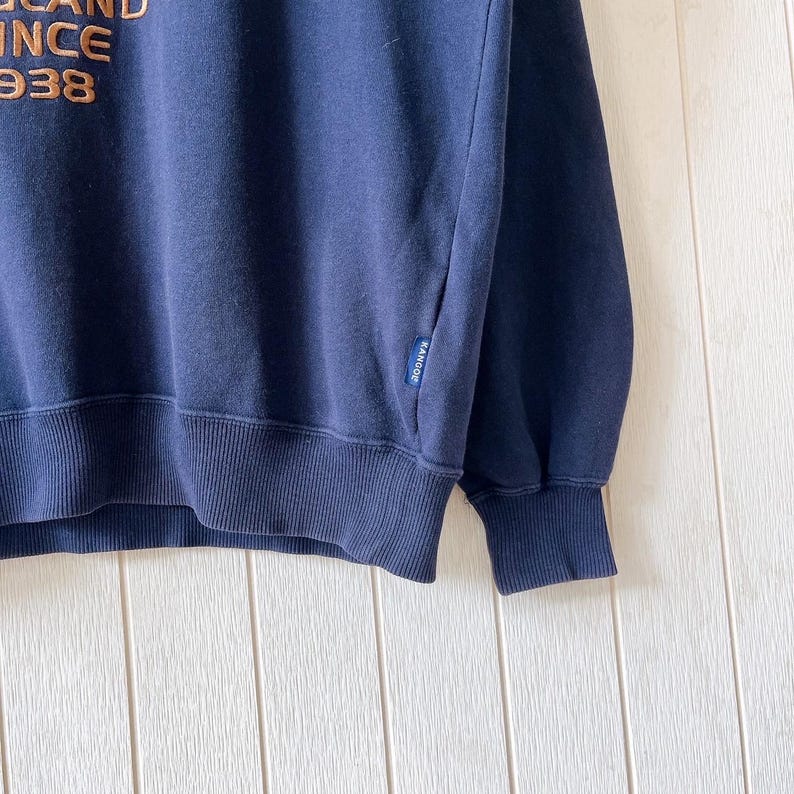 May include: Navy blue sweatshirt with ribbed cuffs and hem. The upper left features the text "BRAND SINCE 1938" in a gold colour. A small tag with the brand name is visible on the sleeve.