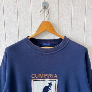 May include: Navy blue sweatshirt featuring the word "CUMBRIA" and a kangaroo logo. The sweatshirt has a crew neck and long sleeves. The brand name "KANGOL" is embroidered on the collar.