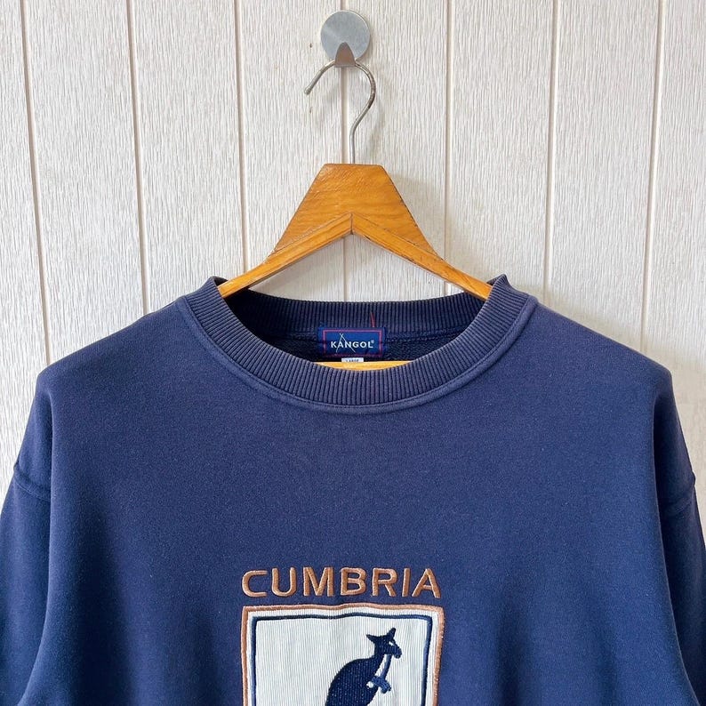 May include: Navy blue sweatshirt featuring the word "CUMBRIA" and a kangaroo logo. The sweatshirt has a crew neck and long sleeves. The brand name "KANGOL" is embroidered on the collar.