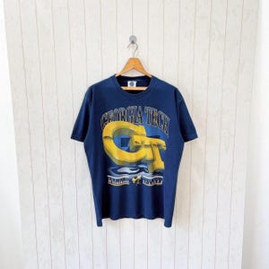 Könnte beinhalten: Marineblaues T-Shirt mit einem großen, 3D-gelben und blauen "GT"-Grafik und dem Text "Georgia Tech". Das Shirt hat einen Rundhalsausschnitt und kurze Ärmel und hängt an einem Holzbügel.