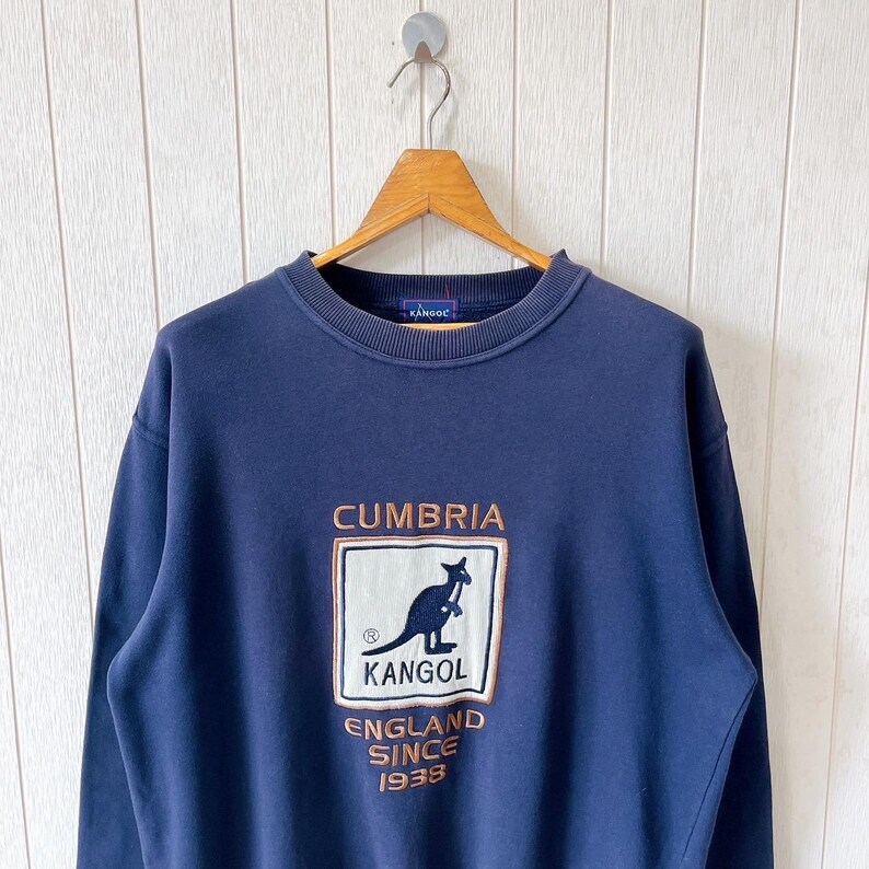 May include: Navy blue sweatshirt featuring the word "CUMBRIA" embroidered in brown above a kangaroo logo. The logo is inside a white rectangle with the word "KANGOL" below the kangaroo. The text "ENGLAND SINCE 1938" is below the logo.