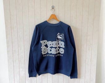 May include: Navy blue crewneck sweatshirt with the text "Penn State" in gray block letters and a graphic of a Nittany Lion. The words "Nittany Lions" are below the text. The sweatshirt is hanging on a wooden hanger.
