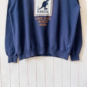 May include: Navy blue sweatshirt with the Kangol logo, a kangaroo emblem in a rectangular frame. The text "KANGOL ENGLAND SINCE 1938" is printed below the logo in gold. The sweatshirt has ribbed cuffs and hem, a classic design.