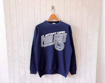 May include: A navy blue crew neck sweatshirt with the text "Nittany Lions Penn State" in white and grey. The sweatshirt also features a lion's head graphic. The garment is suspended from a wooden hanger.