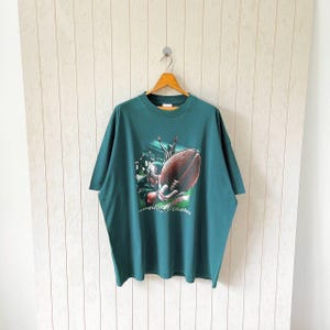 Könnte beinhalten: Grünes T-Shirt mit einem Football-Motiv. Die Grafik zeigt einen Spieler mit einem Football und den Schriftzug "mighty eagles". Das Shirt hängt an einem Holzbügel.