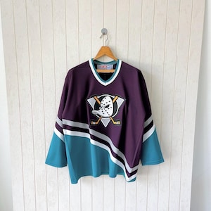 Op de afbeelding: Een paars en teal hockey shirt met een wit en zilver streepontwerp. Het shirt heeft een V-hals en een logo van een eendenmasker karakter met gekruiste hockeysticks. Het shirt hangt aan een houten hanger.