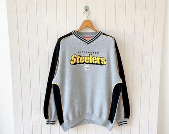 Puede incluir: Sudadera gris de los Steelers de Pittsburgh con mangas negras y detalles amarillos. La palabra "Steelers" es amarilla con contorno negro, y "Pittsburgh" está arriba. El cuello en V tiene rayas negras y blancas.