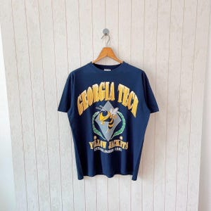 Könnte beinhalten: Marineblaues T-Shirt mit einem gold-gelben Grafik einer Biene und den Worten "GEORGIA TECH" und "YELLOW JACKETS". Das T-Shirt hängt an einem Holzbügel vor einer weißen Holzvertäfelung.