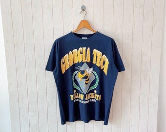 May include: Navy blue t-shirt with a gold and yellow graphic of a bee and the words "GEORGIA TECH" and "YELLOW JACKETS". The shirt is hanging on a wooden hanger against a white wood-paneled wall.