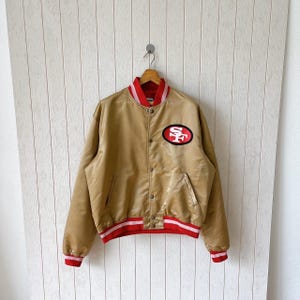 May include: A gold satin bomber jacket featuring a red and white striped collar, cuffs, and waistband. The front displays a black oval patch with the white and red letters SF. The jacket has a snap-button closure and two side pockets.