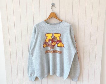 May include: A gray sweatshirt with a large gold and maroon "M" logo and a cartoon beaver mascot. The word "MINNESOTA" is below the logo. The right sleeve has vertical text in maroon. The sweatshirt is hanging on a wooden hanger.