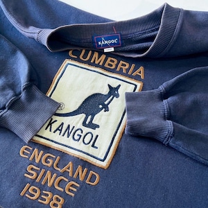 May include: A navy blue Kangol sweatshirt with the brand's logo, a kangaroo, and the text "Cumbria England Since 1938". It has a crew neck and ribbed cuffs. A Kangol label is sewn on the back of the neck.