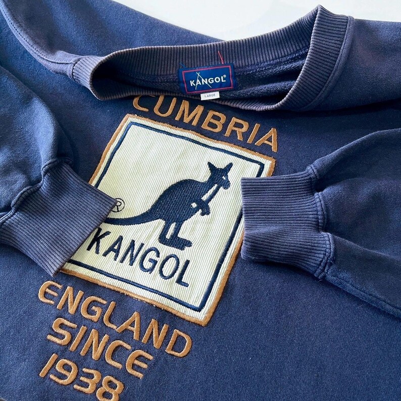 May include: A navy blue Kangol sweatshirt with the brand's logo, a kangaroo, and the text "Cumbria England Since 1938". It has a crew neck and ribbed cuffs. A Kangol label is sewn on the back of the neck.